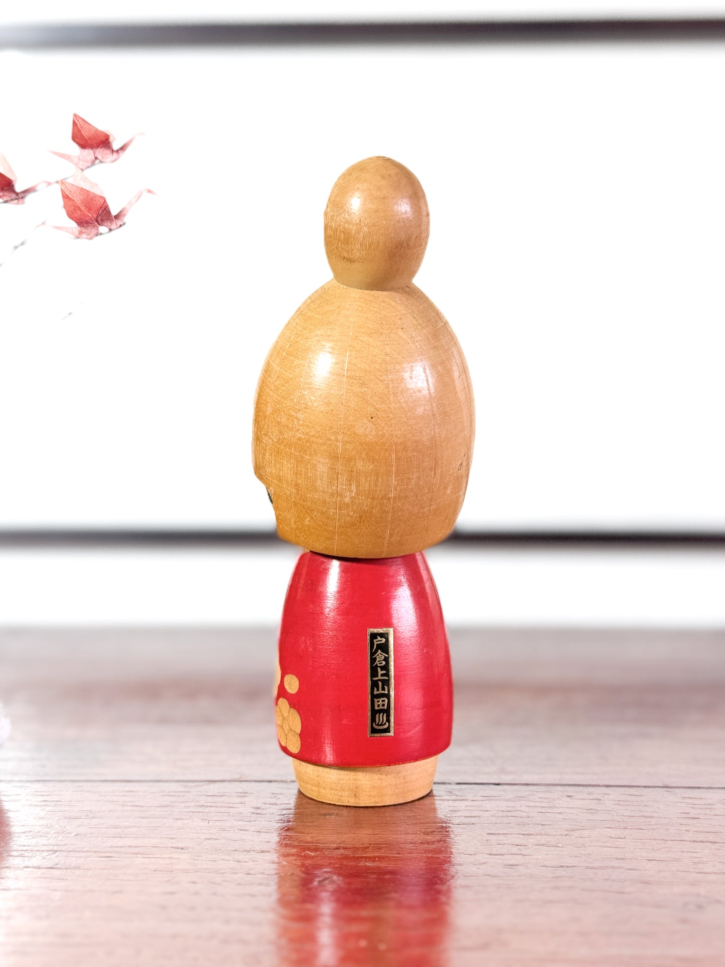 kokeshi, poupée japonaise en bois de Sato Suigai, avec tête penchée, visage souriant et kimono rouge avec fleurs de cerisier gravées, vu de dos