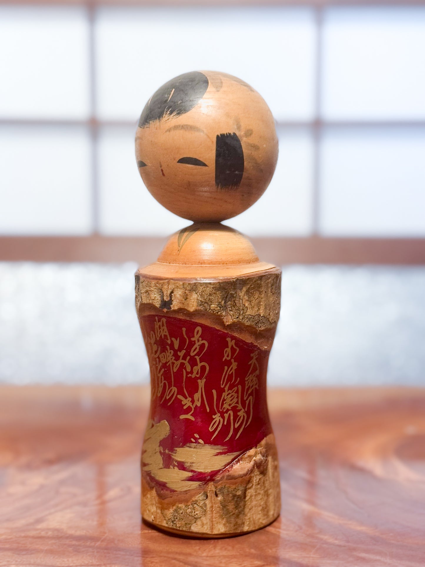 Kokeshi en bois brut avec écorce et laque rouge, décorée de kanji dorés, vu de 3/4 gauche