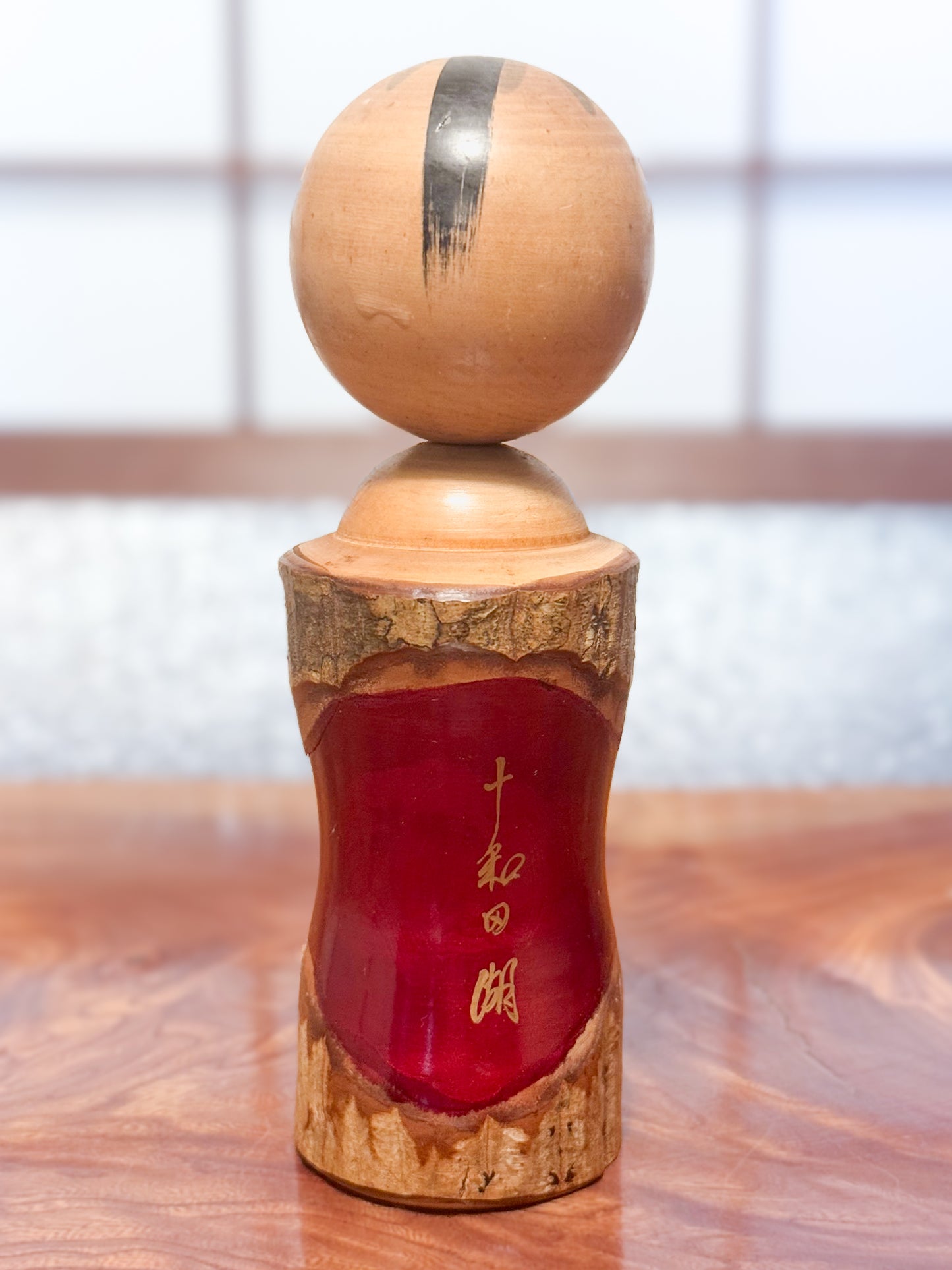 Kokeshi en bois brut avec écorce et laque rouge, décorée de kanji dorés, vu de dos avec la signature de l'artisan
