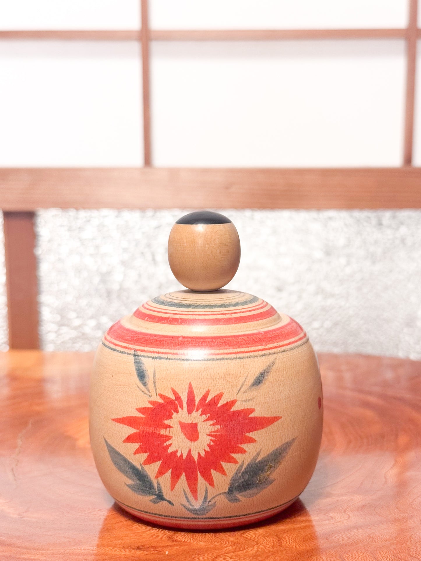 Kokeshi ronde évoquant un bébé dans son panier, décorée de grands chrysanthèmes rouges sur bois clair, vu de dos
