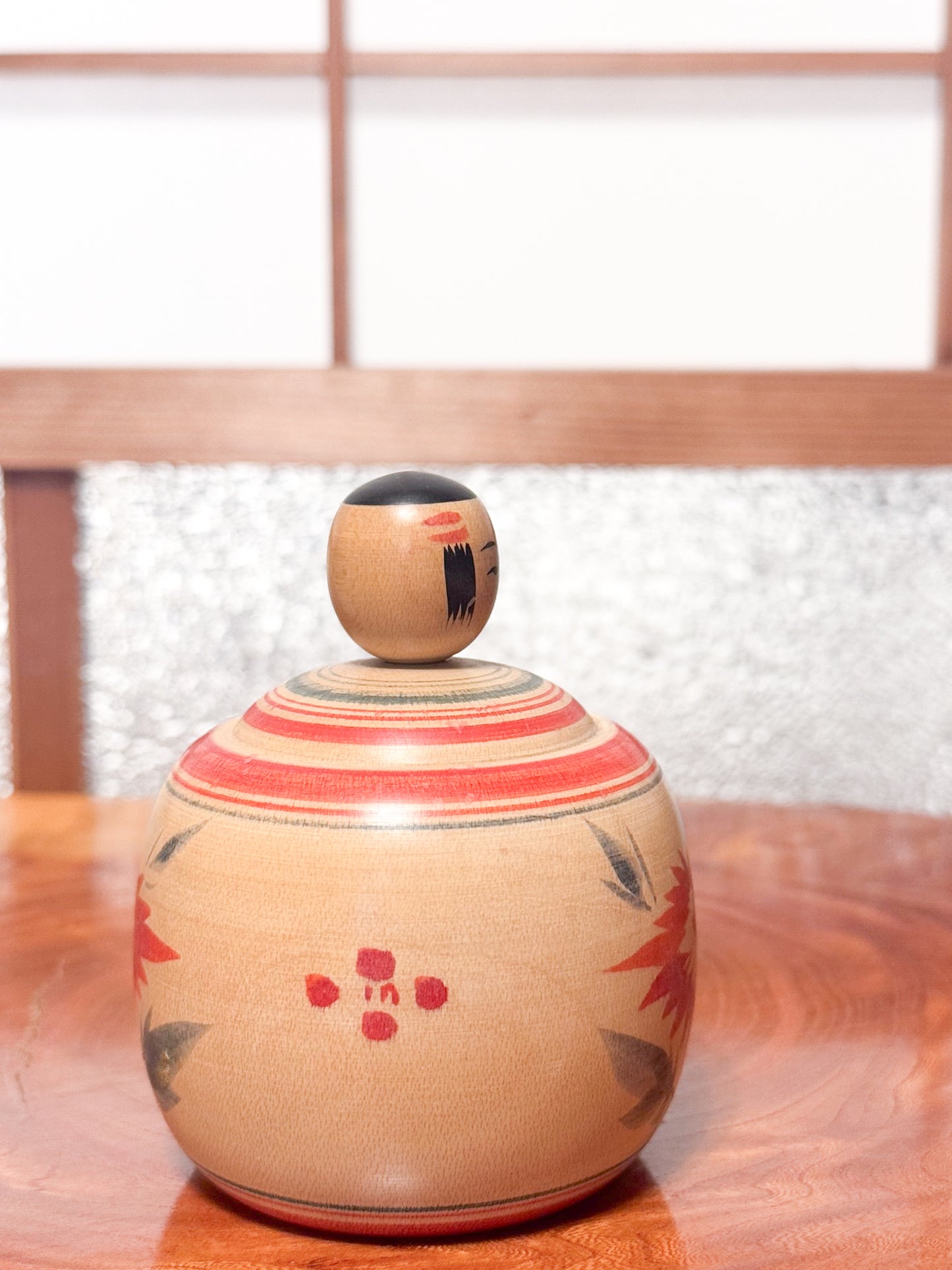 Kokeshi ronde évoquant un bébé dans son panier, décorée de grands chrysanthèmes rouges sur bois clair, profil droit