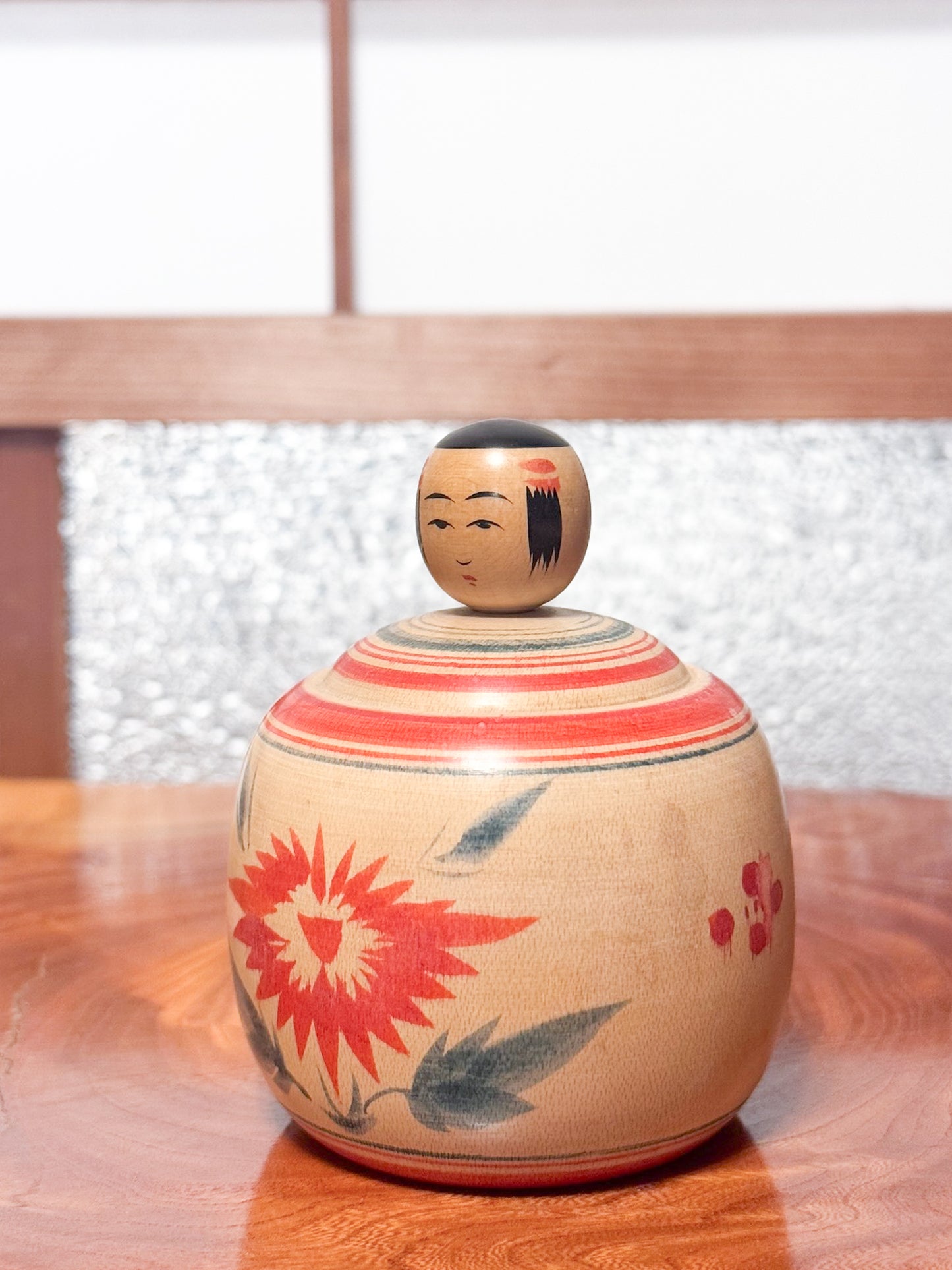 Kokeshi ronde évoquant un bébé dans son panier, décorée de grands chrysanthèmes rouges sur bois clair, vu de 3/4