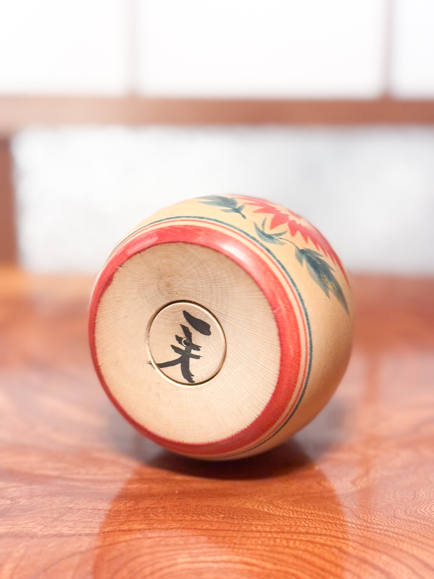 Kokeshi ronde évoquant un bébé dans son panier, décorée de grands chrysanthèmes rouges sur bois clair, dessous la signature de l'artisan