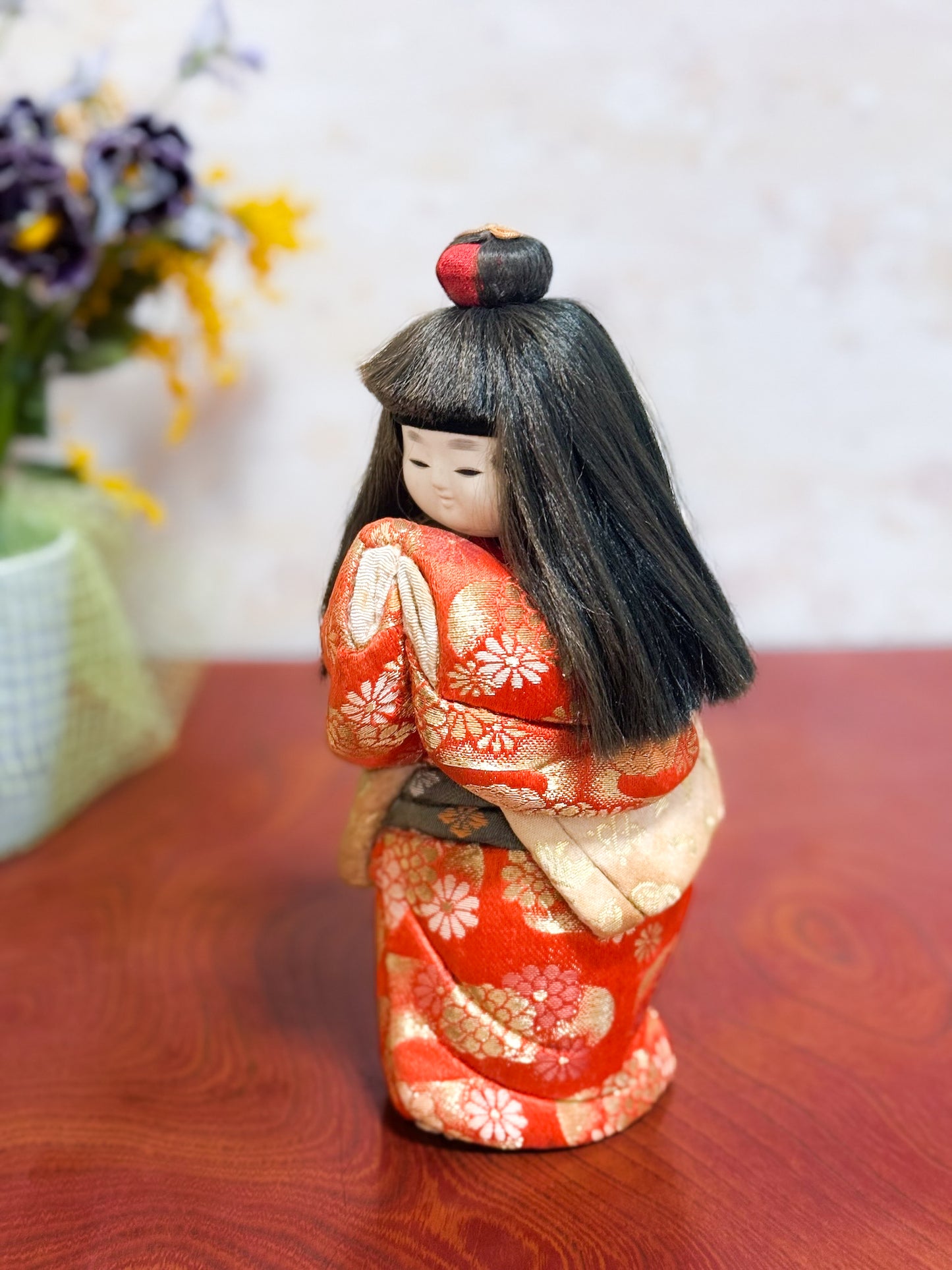 Poupée japonaise kimekomi représentant une jeune fille en kimono rouge fleuri avec les mains jointes, coiffée d’un chignon traditionnel, vu de 3/4