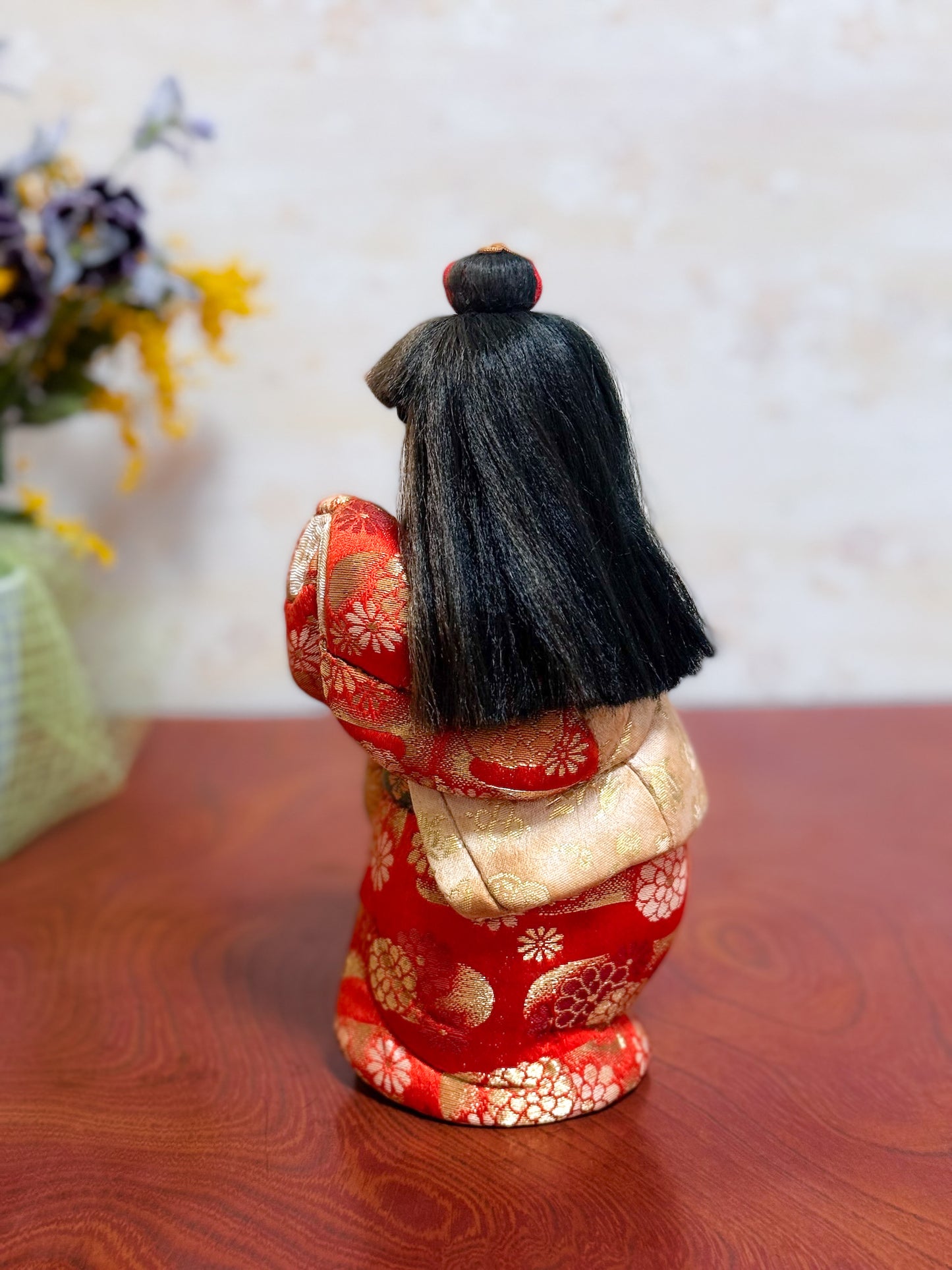 Poupée japonaise kimekomi représentant une jeune fille en kimono rouge fleuri avec les mains jointes, coiffée d’un chignon traditionnel, profil gauche