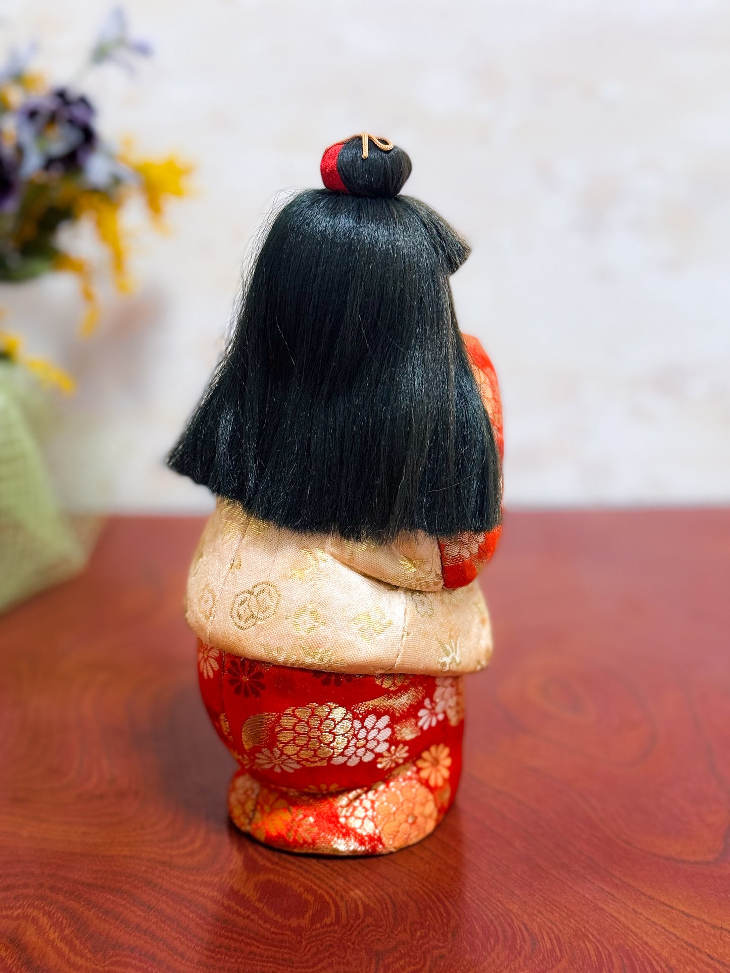 Poupée japonaise kimekomi représentant une jeune fille en kimono rouge fleuri avec les mains jointes, coiffée d’un chignon traditionnel, vu de dos