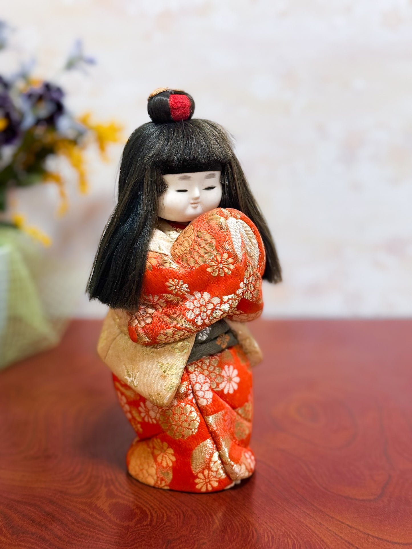 Poupée japonaise kimekomi représentant une jeune fille en kimono rouge fleuri avec les mains jointes, coiffée d’un chignon traditionnel, profil droit