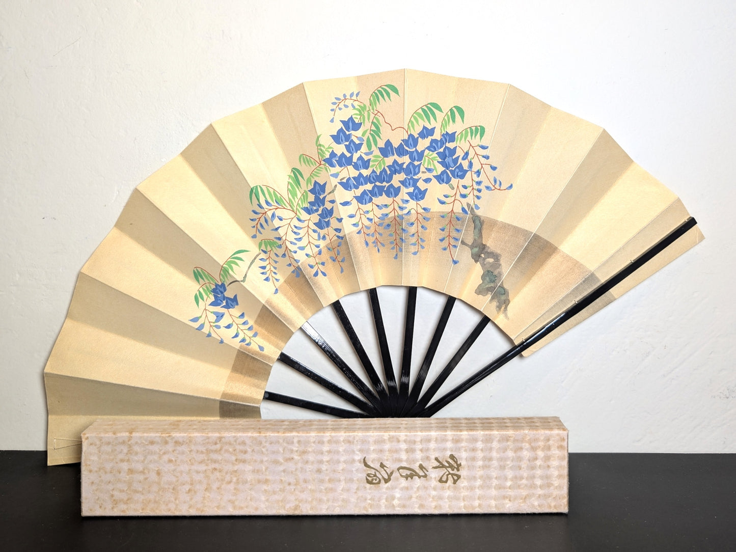 Eventail traditionnel japonais de théâtre kabuki en papier washi écru micacé et bois laqué noir. Peint à la main avec peinture d'une branche de glycine, fleurs bleues feuilles vertes. Avec boite cartonnée, écriture imprimée type calligraphie dorée.
