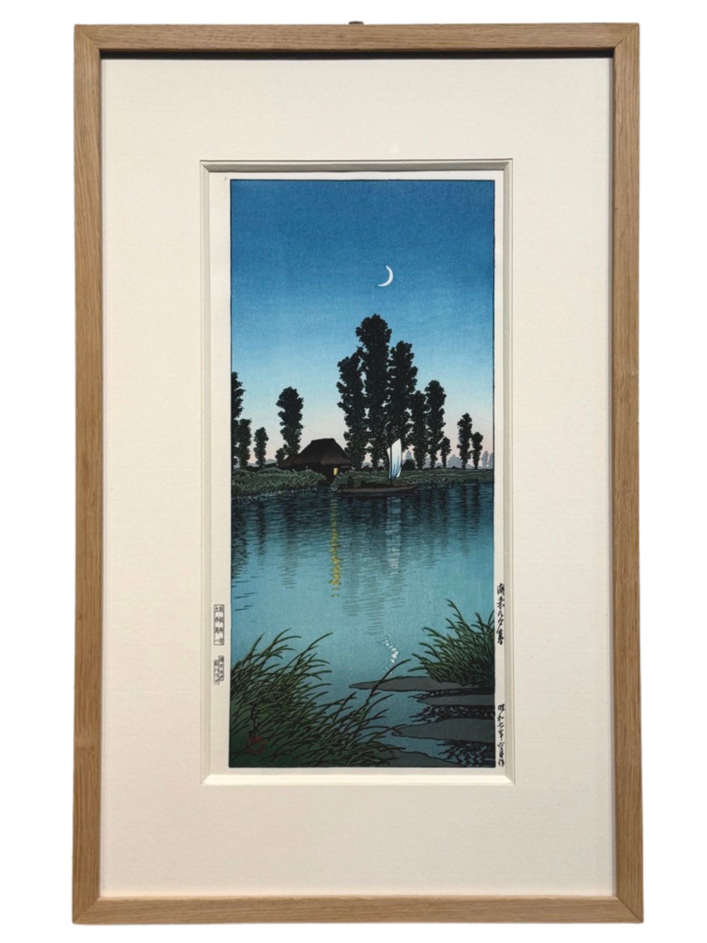 estampe japonaise encadrée de Hasui Kawase, d'un paysage nocturne avec un croissant de lune, une rivière, un bateaux à voile passant devant une chaumière éclairée, en format long et étroit