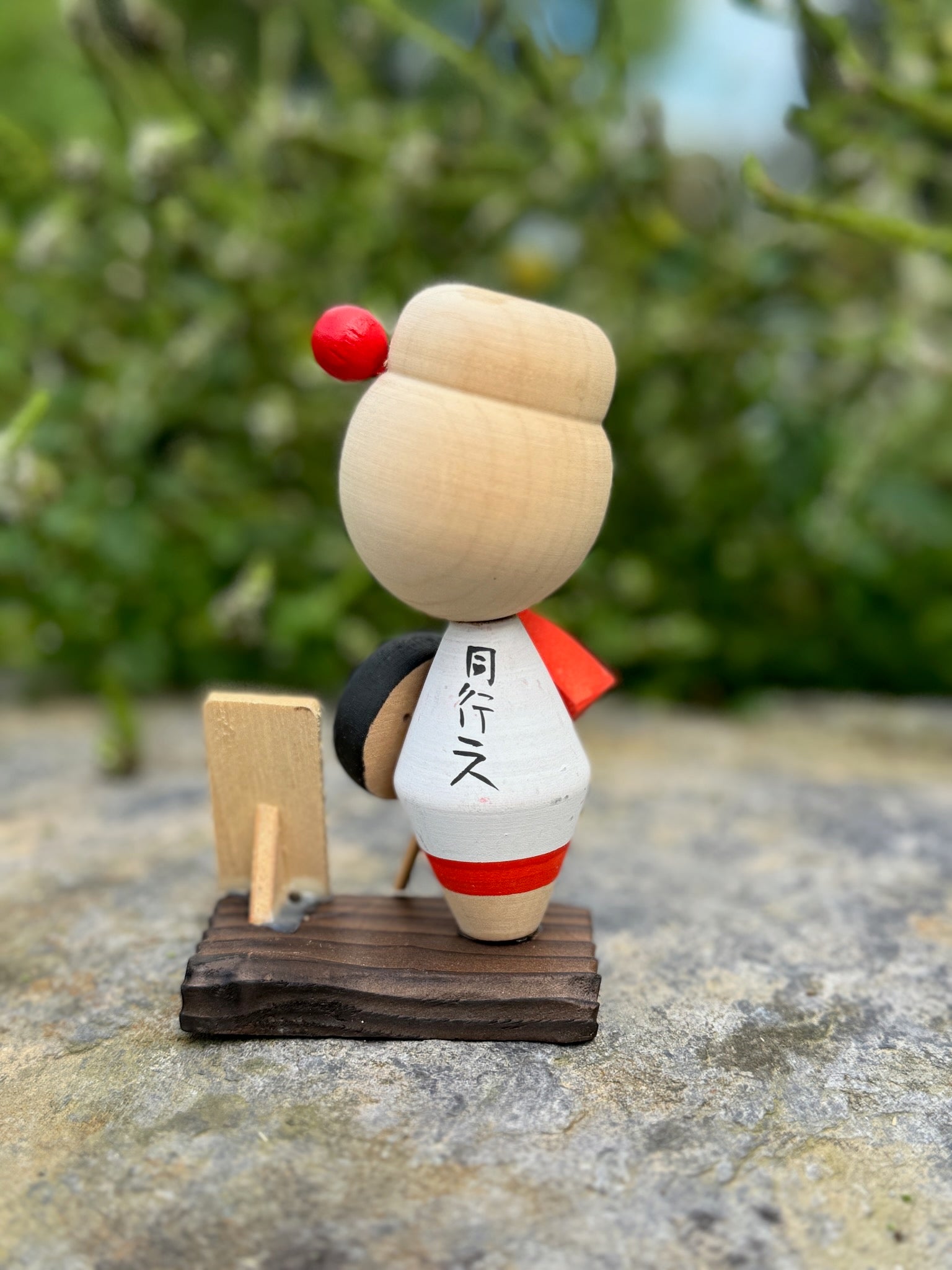 Kokeshi souvenir, poupée en bois japonaise, kimono orange et chapeau noir et blanc, pèlerinage de shikoku, épingle à cheveux rouge ronde, ruban peint et fleurs blanches sur chignon. Dos Kokeshi veste blanche et écriture.