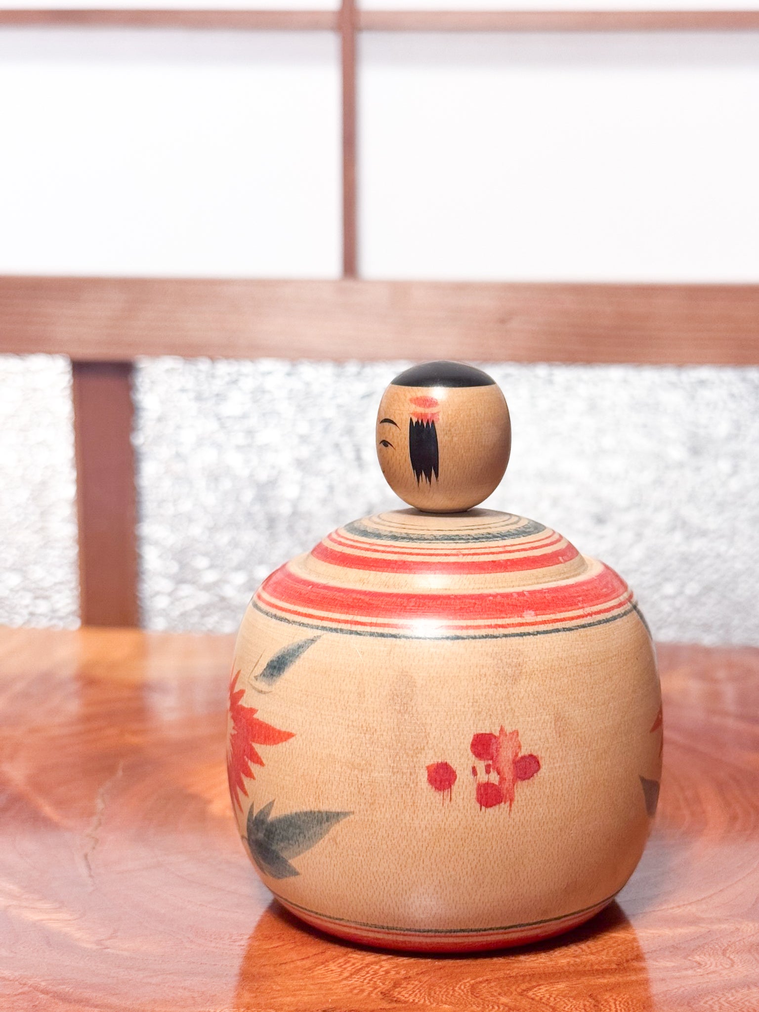 Kokeshi ronde évoquant un bébé dans son panier, décorée de grands chrysanthèmes rouges sur bois clair, profil gauche