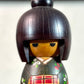 kokeshi gunma de Sadao Kishi au kimono vert, fleurs stylisées et obi rouge à carreaux, gros plan sur le visage souriant