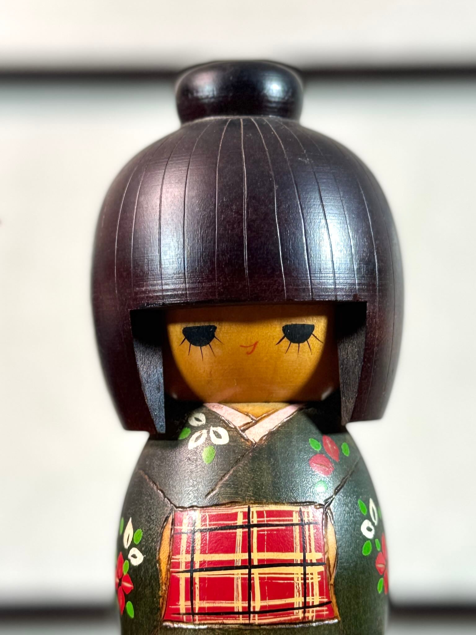 kokeshi gunma de Sadao Kishi au kimono vert, fleurs stylisées et obi rouge à carreaux, gros plan sur le visage souriant