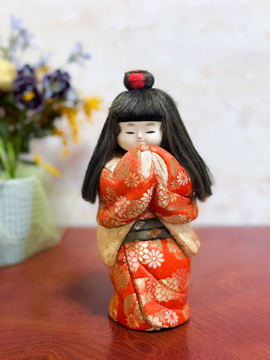 Poupée japonaise kimekomi représentant une jeune fille en kimono rouge fleuri avec les mains jointes, coiffée d’un chignon traditionnel, vu de face