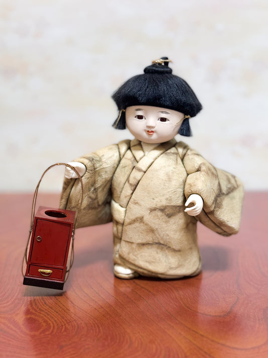 Poupée japonaise kimekomi représentant un enfant en kimono beige tenant une boîte à tabac rouge