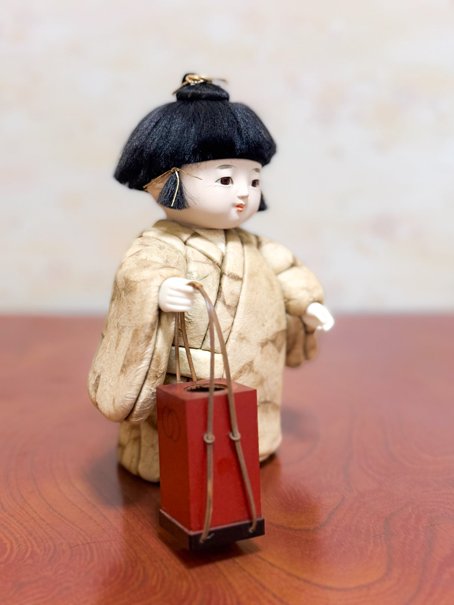 Poupée japonaise kimekomi représentant un enfant en kimono beige tenant une boîte à tabac rouge, profil droit