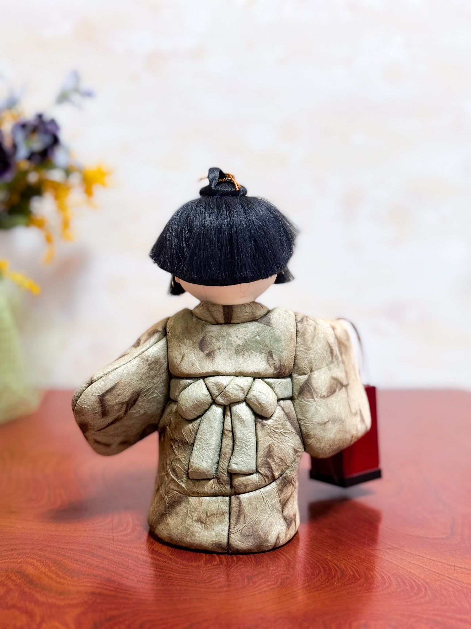 Poupée japonaise kimekomi représentant un enfant en kimono beige tenant une boîte à tabac rouge, vu de dos
