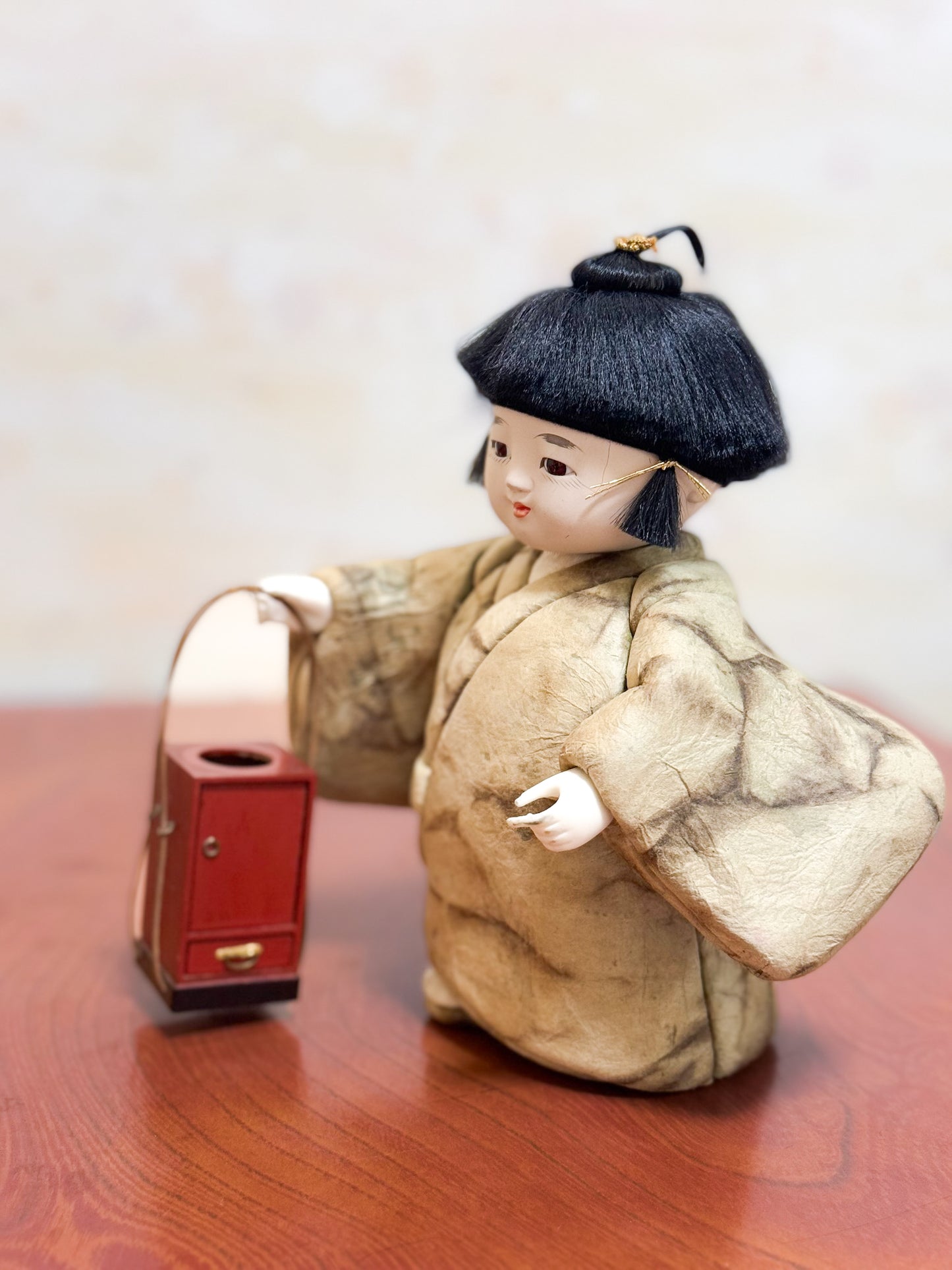 Poupée japonaise kimekomi représentant un enfant en kimono beige tenant une boîte à tabac rouge, profil gauche