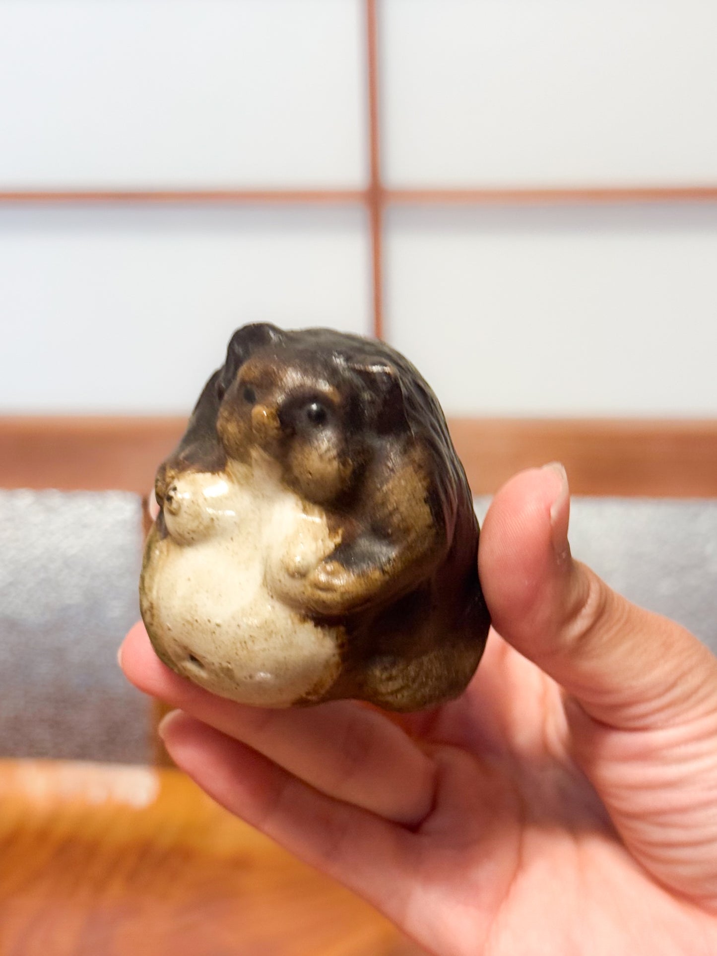 Figurine japonaise en céramique représentant un tanuki assis avec ventre arrondi, émail brun et crème, tenue en main, profil
