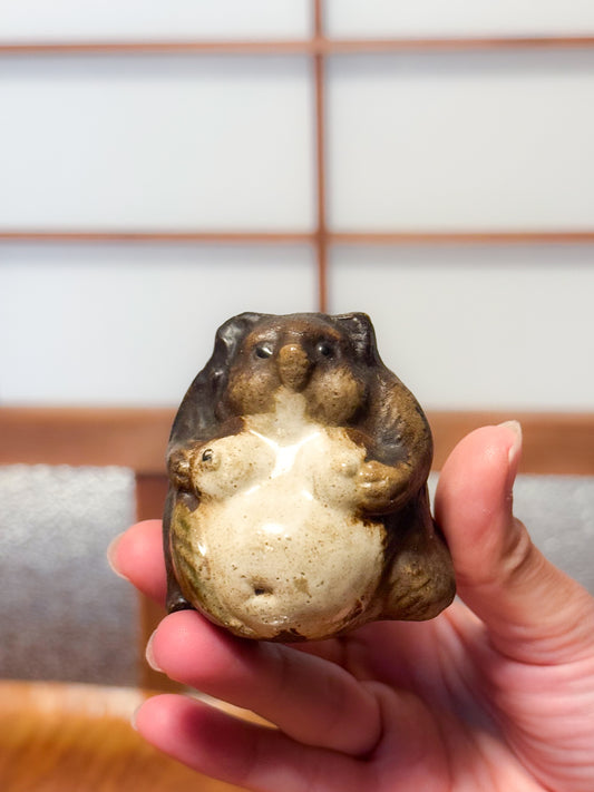 Figurine japonaise en céramique représentant un tanuki assis avec ventre arrondi, émail brun et crème, tenue en main