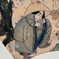 Estampe japonaise en forme d'éventail parsemée de fragments d'or et d'argent, gros plan femme qui boit de l'eau avec une louche accroupie sous un chêne, chapeau, puits.
