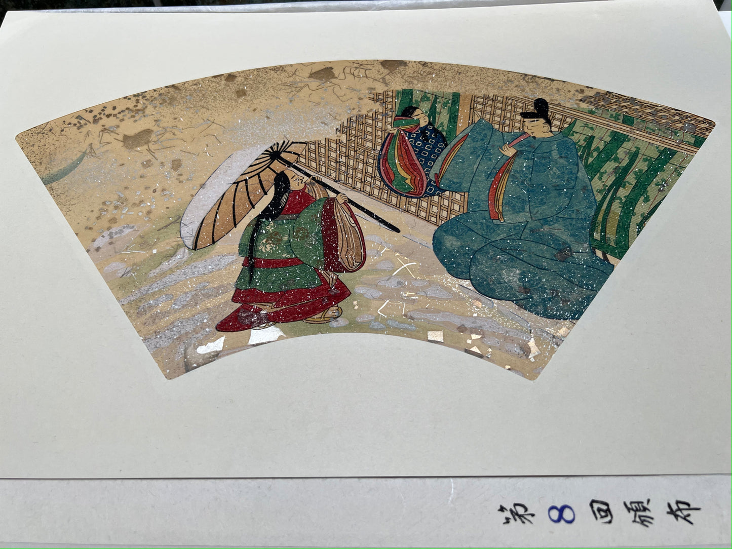 Estampe japonaise en forme d'éventail parsemée de fragments d'or et d'argent, matin neigeux, une vieille femme se protège de la neige sous une ombrelle, un homme tenant un éventail l'accueille devant une maison traditionnelle japonaise, une femme les observe à la fenêtre, série sutra du lotus n°8 écrit en japonais dans le coin inférieur droit.