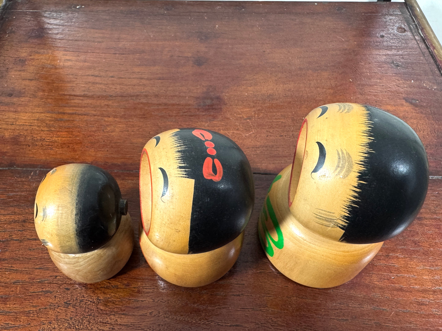 Trio de kokeshi en bois riant aux éclats avec calligraphie rouge et verte de Sato Kouson, vu de dessus, noeud rouge dans les cheveux