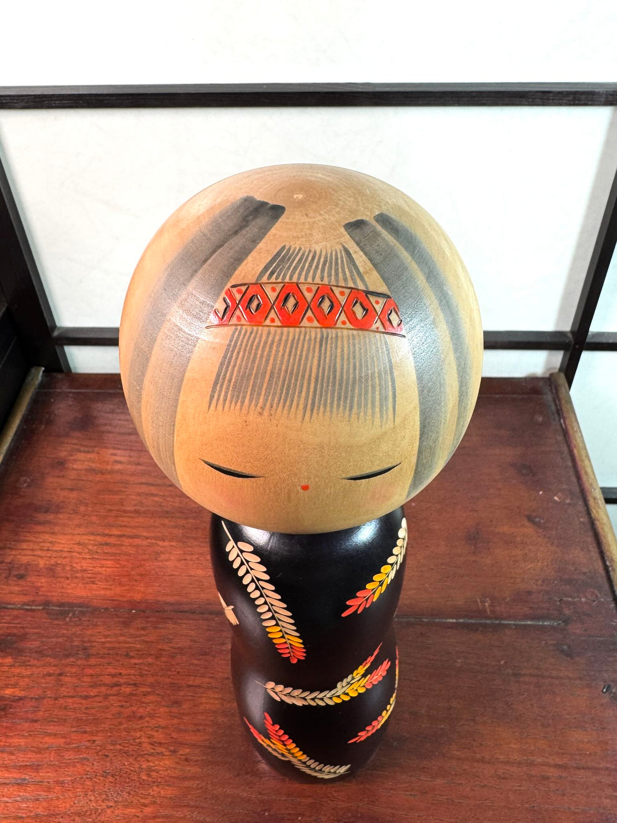 Grande kokeshi vintage de Yuji Kawase au corps noir décoré d’épis de riz beige, jaunes et rouges, Japon, période Shōwa. Le dessus de la tête avec un bandeau aux motifs losanges rouges