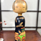 Kokeshi vintage de Yuji Kawase au corps noir décoré d’épis de riz et de deux récipients colorés à motifs géométriques.