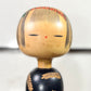 Kokeshi vintage de Yuji Kawase au corps noir décoré d’épis de riz et de deux récipients colorés à motifs géométriques, gros plan sur le visage