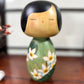 Kokeshi vintage de Yuji Kawase datée de 1994, au corps vert décoré de fleurs blanches et à la coiffure noire asymétrique, portrait