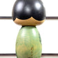 Kokeshi vintage de Yuji Kawase datée de 1994, au corps vert décoré de fleurs blanches et à la coiffure noire asymétrique, la signature et la date gravées au dos