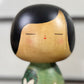 Kokeshi vintage de Yuji Kawase datée de 1994, au corps vert décoré de fleurs blanches et à la coiffure noire asymétrique, gros plan sur le visage souriant