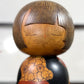 Kokeshi vintage de Yūji Kawase datée de 1977, en bois, à fleurs pyrogravées, grands aplats noirs et rouge brique en puzzle et visage aux yeux clos, gros plan sur le visage