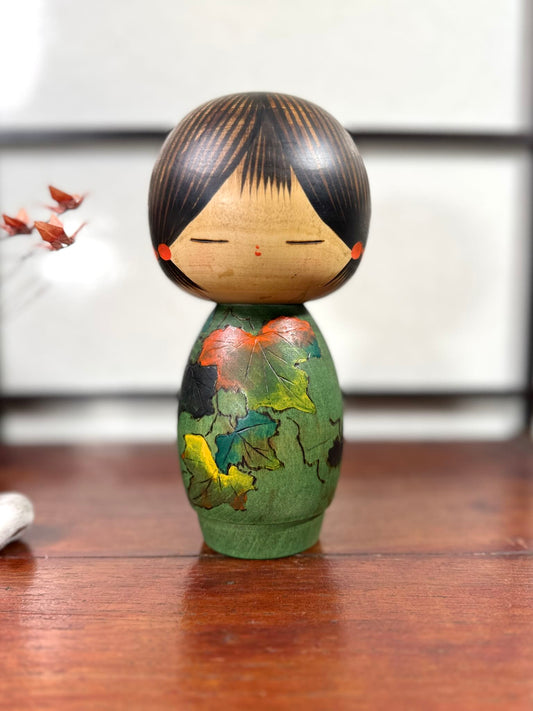 Kokeshi vintage de Yūji Kawase datée de 1981, corps vert décoré de feuilles rouges, jaunes et vertes, tête ronde aux cheveux noirs striés et yeux clos, 20 cm.