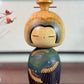 Kokeshi Yuji Kawase bleu-vert avec parapluies gravés et chignon.