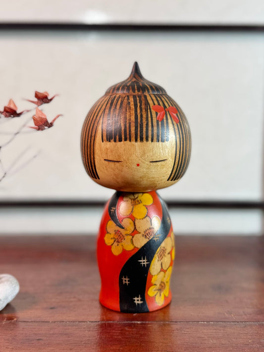 Kokeshi Yuji Kawase au kimono rouge fleuri et coiffure pointue.