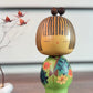 Kokeshi Yuji Kawase au kimono vert fleuri et coiffure à boules.