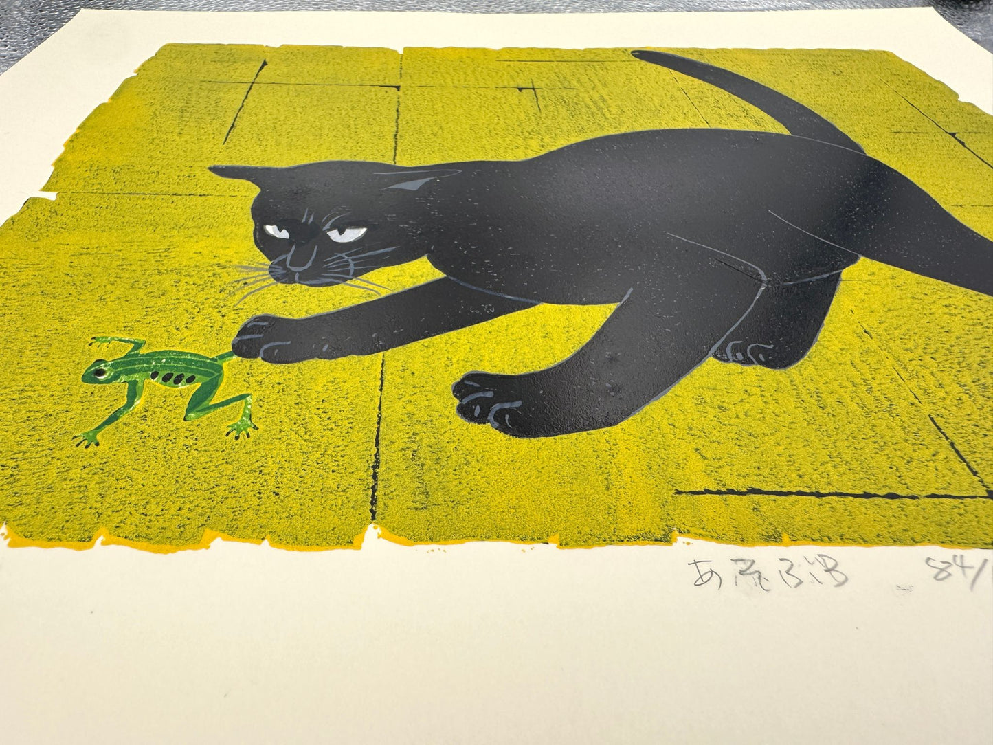 Estampe japonaise de Nishida montrant un chat noir jouant avec une grenouille sur fond vert et jaune, vue de profil pour le détail du fond
