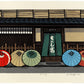 estampe japonaise contemporaine de Nishijima  entrée d'une maison traditionnelle japonaise de facade en bois avec noren. Cinq parapluies colorés