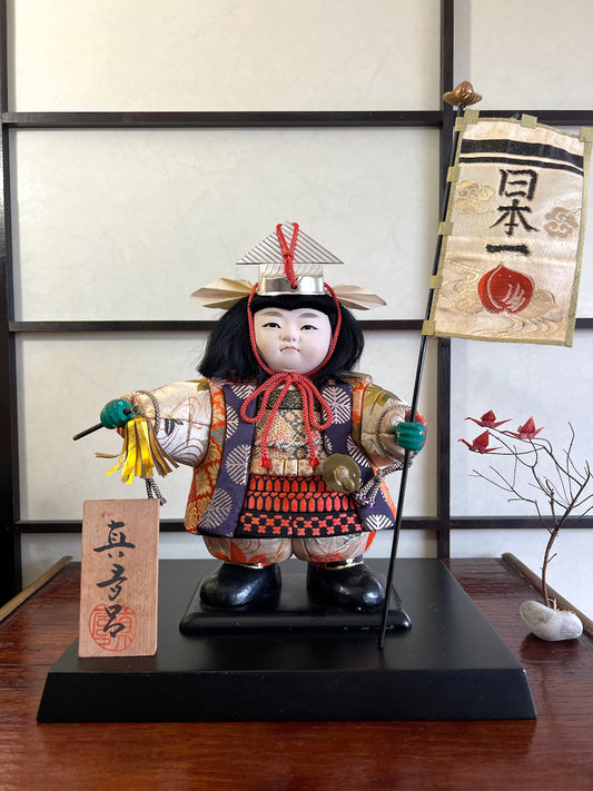 poupée traditionnelle kimekomi kintaro en samurai portant drapeau et fouet sur socle