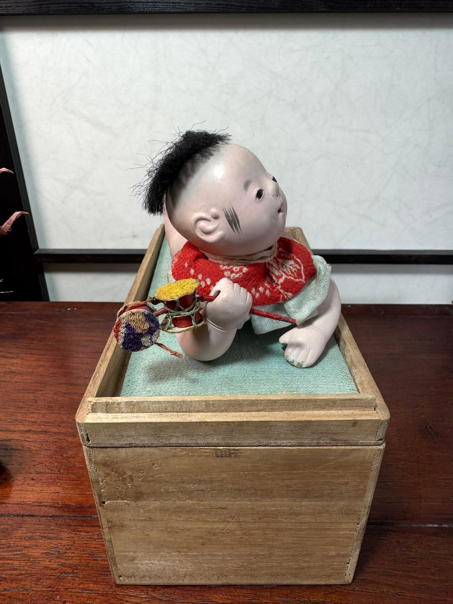 Poupée japonaise ancienne bébé couché en terre cuite sur boite en bois, avec un hochet à la main