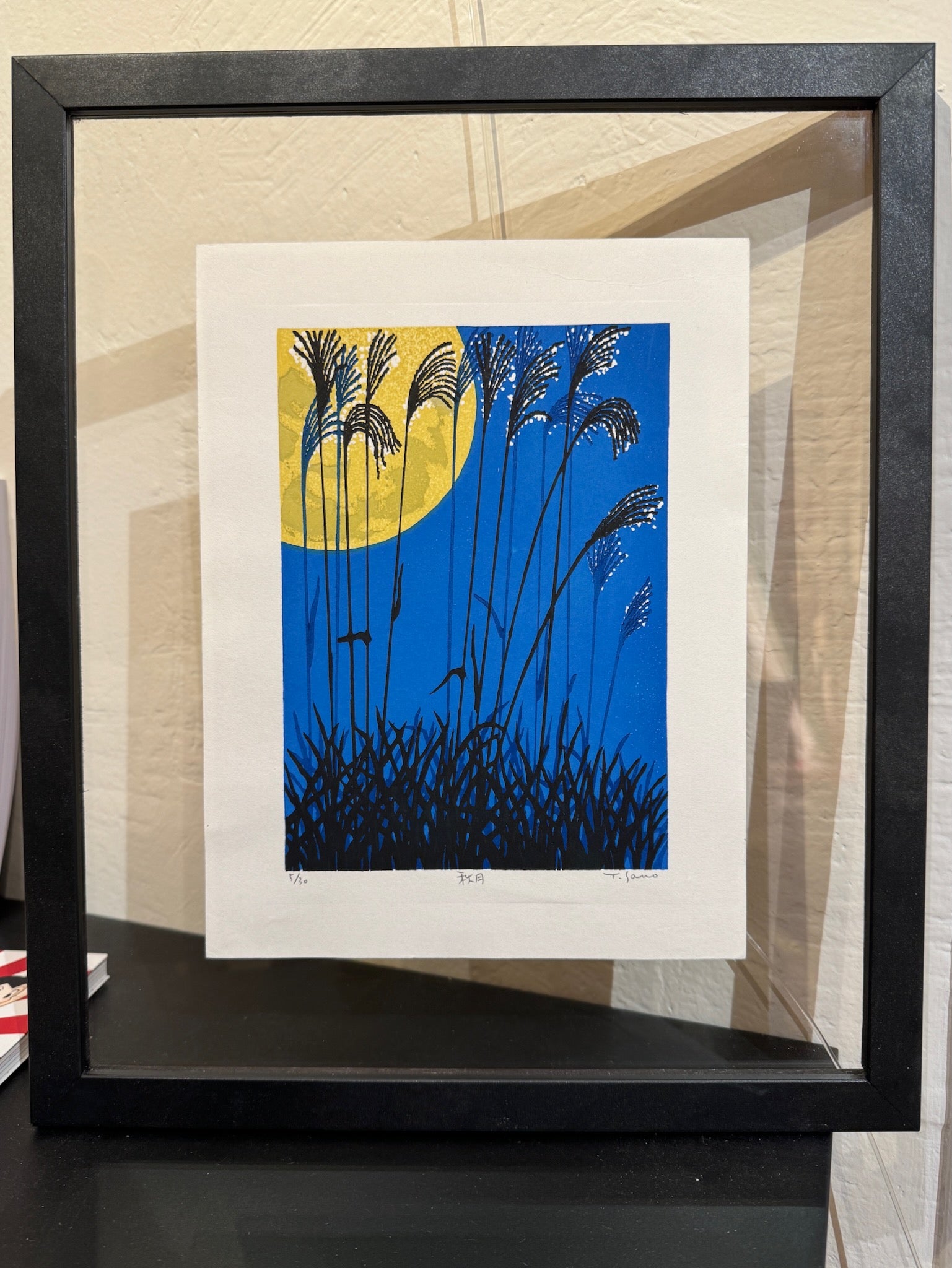 estampe japonaise contemporaine encadrée de Sano Takao, la pleine lune en automne avec herbes graminées en premier plan, entre-deux-verre et cadre noir, posé sur un meuble
